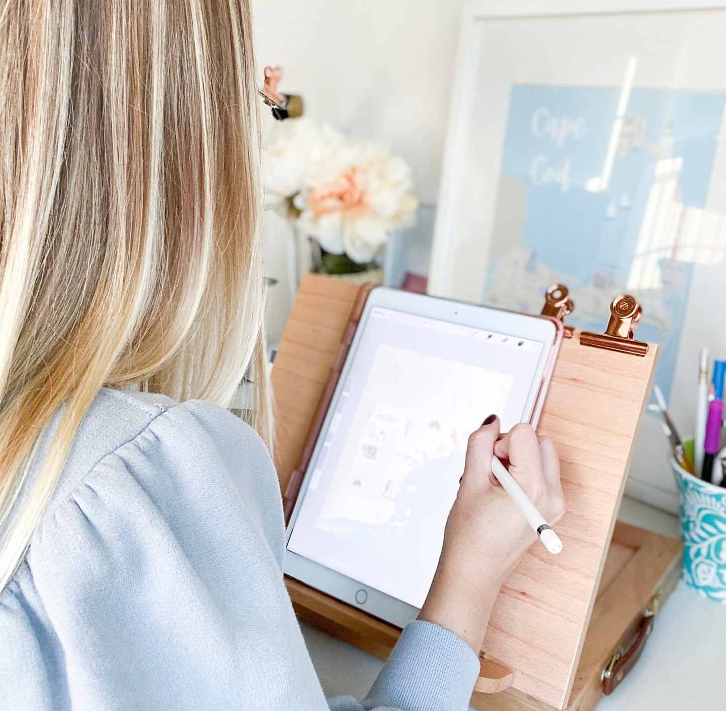 A woman with blonde hair, seen from behind, uses a white stylus on a tablet placed on a light wood desktop stand with gold-toned clips in a bright, modern home office setting.
