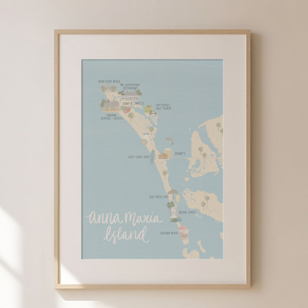 A framed art print of Anna Maria Island, New Mexico, featuring a light blue map with beige landmasses and white script text, displayed on a warm beige wall with soft window shadow patterns.